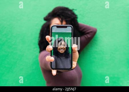 Hands of woman showing selfie on smart phone in front of green wall Stock Photo