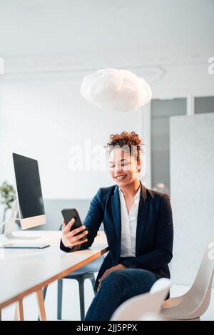 Cloud levitating over happy businesswoman using smart phone in office Stock Photo