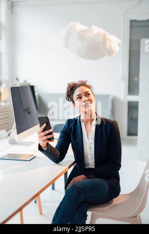 Smiling young businesswoman holding mobile phone looking at levitating cloud in office Stock Photo