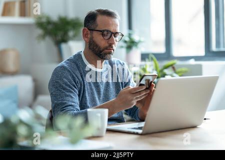 Freelancer using smart phone sitting with laptop at home Stock Photo