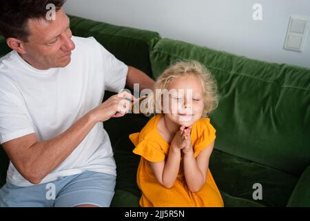 Smiling girl with eyes closed sitting on sofa by father combing her hair at home Stock Photo
