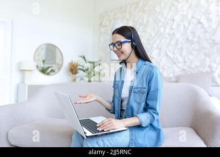 Beautiful woman studying at home, talking on video call with teacher, using headset, woman working sitting on sofa in bright living room Stock Photo