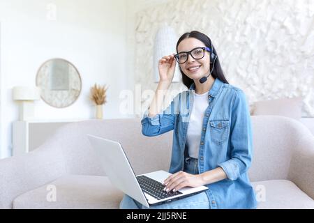 Beautiful young business woman working remotely at home, sitting on sofa and using laptop and headset for video call, woman in glasses smiling and looking at camera Stock Photo