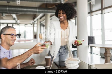 Happy young businesswoman giving salad box to businessman sitting at desk Stock Photo