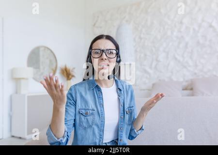 Beautiful woman looking at web camera, nervous and arguing with interlocutor, using headset for video call, working at home sitting on sofa, online conference and study Stock Photo
