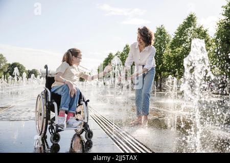 Girl in wheelchair playing along fountain water with mother at park Stock Photo