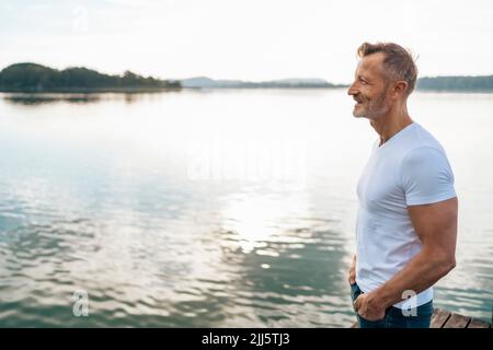 Smiling mature man with hands in pockets standing at lake Stock Photo