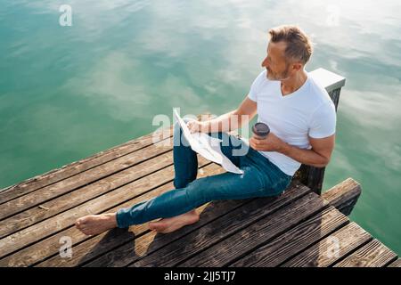 Man with newspaper and disposable coffee cup sitting on pier Stock Photo