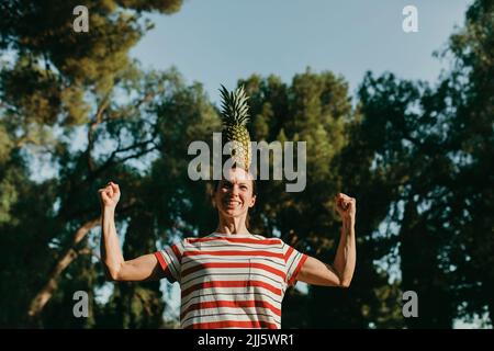 Woman balancing pineapple on head and flexing muscles in park Stock Photo