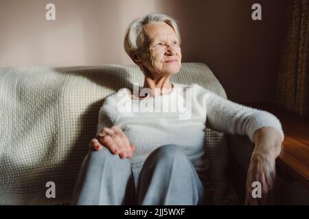 Smiling senior woman with white hair sitting on sofa at home Stock Photo