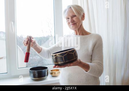 Happy senior woman with singing bowl by window at home Stock Photo