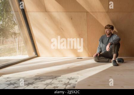 Man sitting on the floor in wooden eco house looking out of window Stock Photo