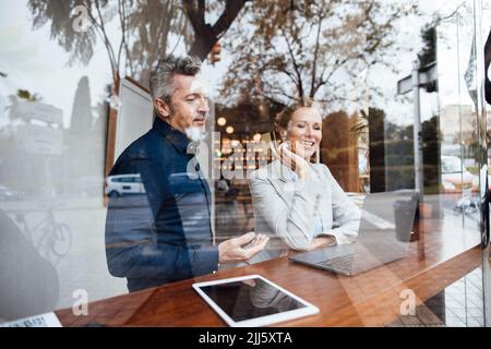 Businesswoman holding credit card sitting by businessman looking at laptop seen through glass Stock Photo