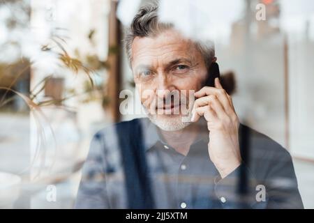 Businessman talking on mobile phone in cafe seen through glass Stock Photo