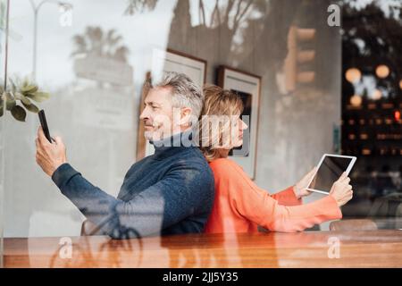 Smiling couple using wireless technologies sitting cafe seen through glass Stock Photo