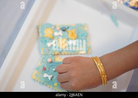 Close up on kid's hands. happy little kid decorating cookies in the tray at school with blue and small fish decorating biscuits and blue sprinkles. Un Stock Photo