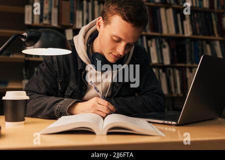 Young man writing while sitting at desk with laptop in library Stock Photo