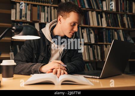 Male student in library looking at laptop. Young man sitting at desk preparing exams at evening. Stock Photo