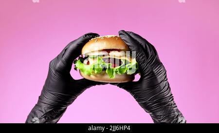 Hands in black latex gloves with burger. Woman holding hamburger. Big appetizing patty with meat cutlet, vegetables, cheese. Fast food concept. Pink Stock Photo