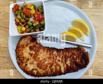 Wiener schnitzel with a bowl of fried bell pepper and onion and three slices of lemon on a white board on a restaurant table Stock Photo