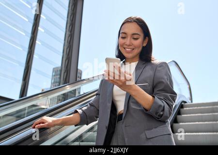 Happy young Asian business woman standing on escalator using phone. Stock Photo