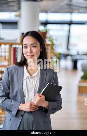 Smiling young Asian business woman standing in office. Vertical portrait Stock Photo