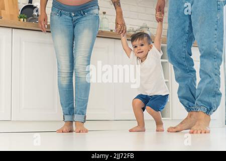 Family of three walking barefoot in their kitchen. Toddler boy standing between his parents and holding their hands in order to jump up. High quality photo Stock Photo