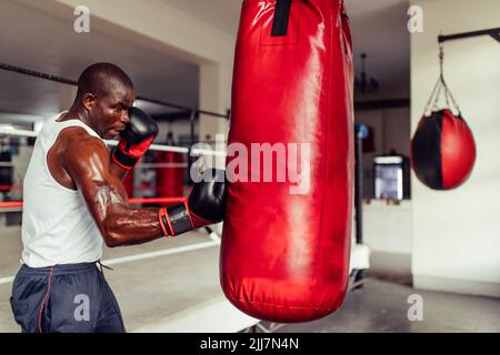 Muscular African boxer practicing his punches on a red leather punching bag. Sport young man having a training session in a boxing gym. Stock Photo