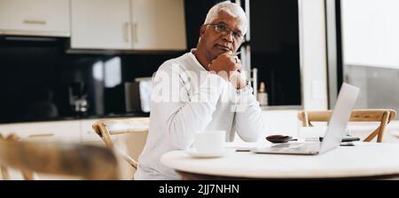 Senior businessman looking at the camera while sitting at a desk in his home. Mature businessman using a laptop while doing remote freelance work. Stock Photo