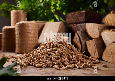 Stack of wood pellets arranged on table near chopped woodpile and compressed biomass briquettes against lush green tree in daytime Stock Photo