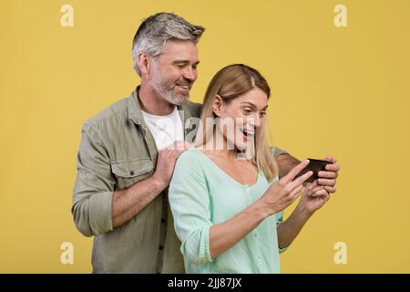 Excited middle aged spouses looking at cellphone screen, using new application, yellow studio background Stock Photo
