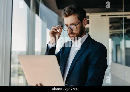 Focused businessman looking at laptop computer, working in modern office, reading emails from his business partners Stock Photo