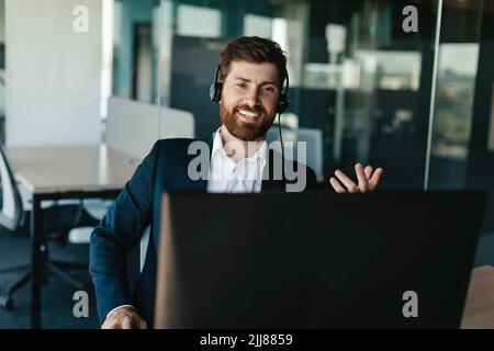 Positive entrepreneur with beard in suit wearing headphones and gesticulating at computer webcamera in office Stock Photo