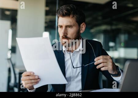 Focused businessman reading negative bills, making business accounting reports, sitting in office interior, copy space Stock Photo