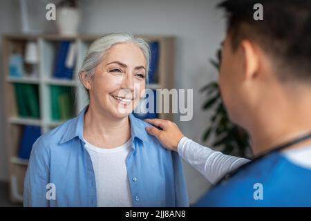 Cheerful senior european lady patient on meeting with young chinese doctor in uniform talking in office Stock Photo