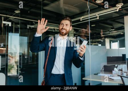 Happy male office worker in suit entering office space smiling and waving to colleagues, copy space Stock Photo