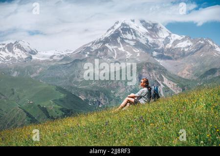 Backpacker woman sitting on a green grass hill and enjoying snowy slopes of Kazbek 5054m mountain with a backpack while she walking by green grass hill Stock Photo