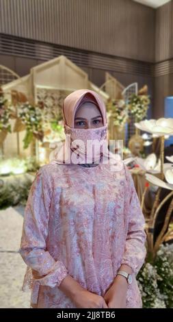 Portrait of an Asian muslim woman looking at camera wearing pink dress, hijab and decorated face mask at a wedding ceremony with bokeh background. Stock Photo