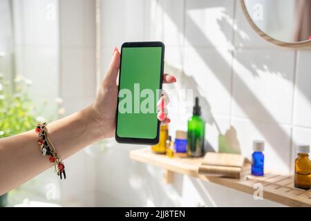 Female hand holding smartphone with green screen in bathroom against bamboo shelf with glass cosmetic bottles. Shadows from the sun on the white tiled wall Stock Photo