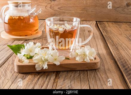glass transparent cup with fragrant jasmine tea on a wooden tray with the flowers of the plant. wooden rustic table with teapot. tea composition Stock Photo
