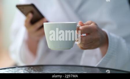 Cropped photo of a woman in a terry bathrobe holding mobile phone and cup Stock Photo
