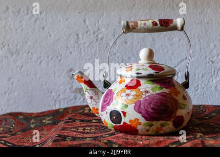 Old teapot decorated with hand-painted flowers, on an old table, with an embroidered mandel, on a white wall Stock Photo