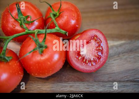 Red tomatoes on vine in kitchen - sliced and whole fresh tomato on wood cutting board - ripe Solanum lycopersicum bunch and cluster Stock Photo