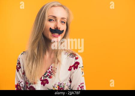 Movember Concept. Copy space, studio shot, isolated on orange background. Funky beautiful caucasian girl with blond hair in a white floral blouse posing to camera with fake black mustache under her nose. High quality photo Stock Photo