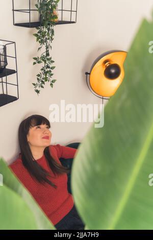 Caucasian brunette woman with bangs smiling sitting on sofa seen through leaves of house plant. Hanging plant. Focus on background. Indoor shot. High quality photo Stock Photo