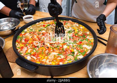 A girl is stirring paella with a black plastic spatula. A large paella bowl on the table. A paella of seafood cocktail, green string beans, red and ye Stock Photo