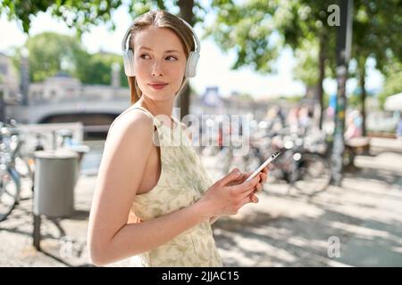 Young pretty woman standing on summer street wearing headphone using smartphone. Stock Photo