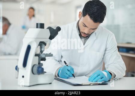 I better record that. a young scientist writing notes while using a microscope in a lab. Stock Photo