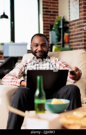 African american man working on laptop and switching tv channels with remote control, browsing internet and finding film to watch. Eating takeout delivery fast food meal at home. Stock Photo