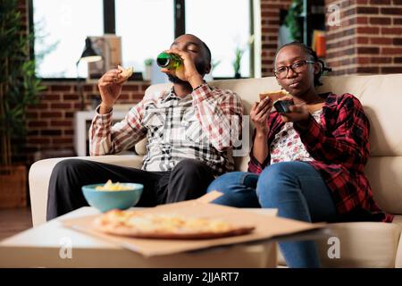 Boyfriend drinking beer from bottle and girlfriend eating pizza while she uses tv remote control to switch channel. Having fun with takeout meal and drink, watching movie on television together. Stock Photo